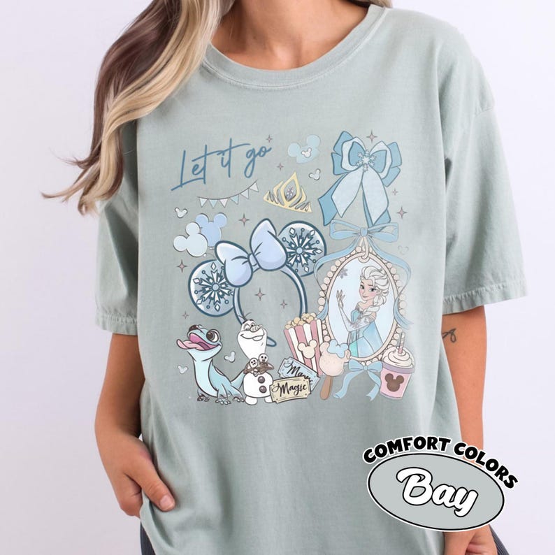 May include: A sage green t-shirt featuring a graphic print with the text Let it go and Magic. The design includes a character from Frozen, a snowman, a lizard, mouse ears with a bow, a tiara, a popcorn bucket, and a drink cup in blue tones.