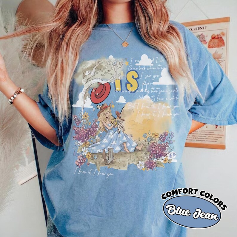 May include: A blue denim-colored t-shirt featuring a graphic print of a blonde character in a blue dress and cowboy boots sitting in a field of purple and white flowers. The design includes a red hat, clouds, and text that reads I know it, I know you.
