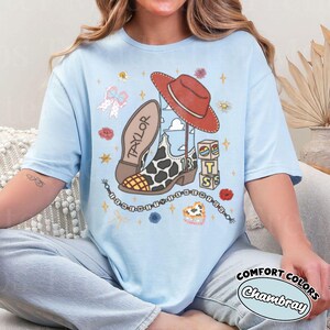 Op de afbeelding: Een lichtblauw t-shirt met een grafische print van een cowboylaars met koeienprint, een rode cowboyhoed, een kralenketting en gestapelde blokken met de tekst "TSS". De laars is gelabeld met "TAYLOR" en "13", met een kralensnoer met "I KNEW YOU".