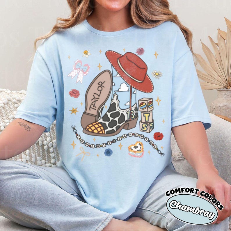 Op de afbeelding: Een lichtblauw t-shirt met een grafische print van een cowboylaars met koeienprint, een rode cowboyhoed, een kralenketting en gestapelde blokken met de tekst "TSS". De laars is gelabeld met "TAYLOR" en "13", met een kralensnoer met "I KNEW YOU".