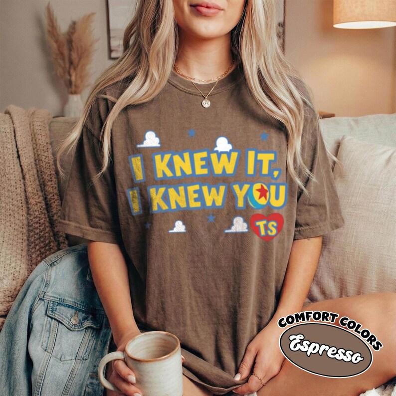 May include: A brown short-sleeved t-shirt featuring the yellow text I KNEW IT, KNEW YOU, a red heart with the letters TS, and small white cloud and blue star graphics. The shirt has a crew neckline and a relaxed fit.