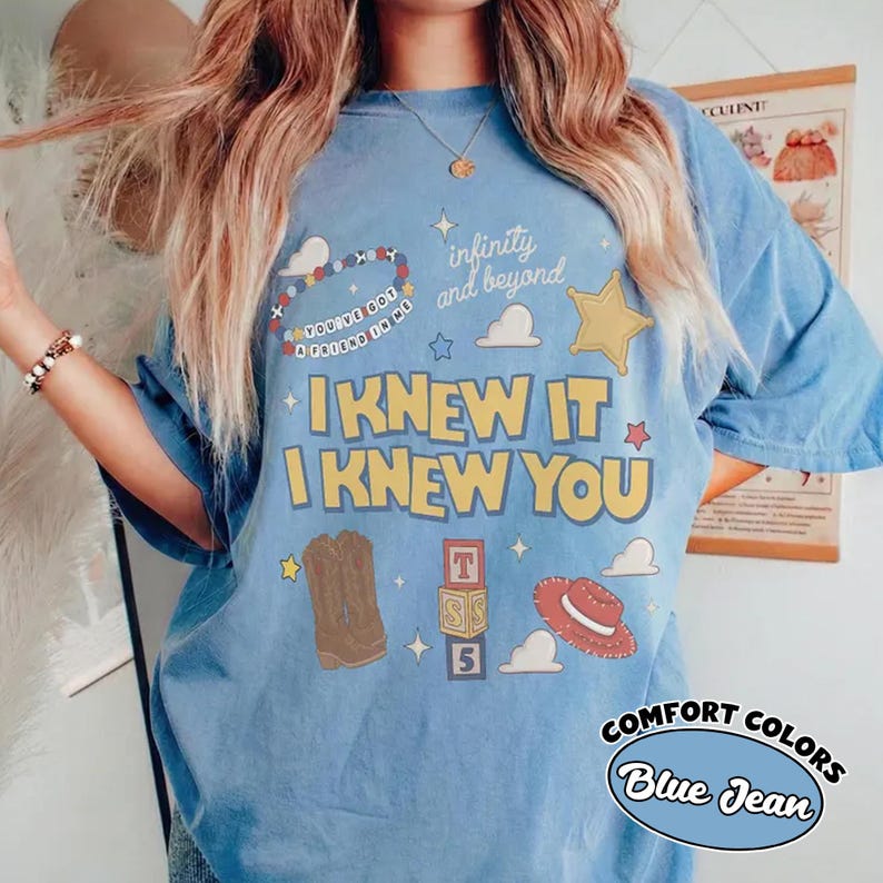 May include: A blue short-sleeve t-shirt featuring a graphic print with the text "I KNEW IT I KNEW YOU", "infinity and beyond", and "YOU'VE GOT A FRIEND IN ME". The design includes illustrations of a beaded bracelet, a star, clouds, cowboy boots, blocks, and a hat.