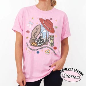 Op de afbeelding: Een lichtroze t-shirt met een grafische print van een cowboylaars met koeienprint, een rode cowboyhoed en een blok met de letters T, S en een zonicoon. De tekst Taylor staat op de laars en eronder hangt een kralensnoer met tekst.