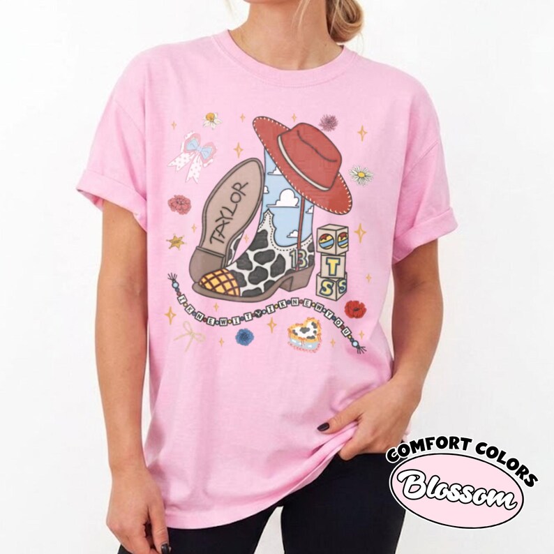 Op de afbeelding: Een lichtroze t-shirt met een grafische print van een cowboylaars met koeienprint, een rode cowboyhoed en een blok met de letters T, S en een zonicoon. De tekst Taylor staat op de laars en eronder hangt een kralensnoer met tekst.
