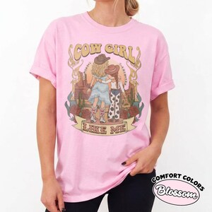 May include: A light pink t-shirt featuring a graphic print of two figures in cowboy hats and boots standing in a desert landscape with cacti and roses. The text COW GIRL and LIKE ME is printed in a decorative font above and below the illustration.