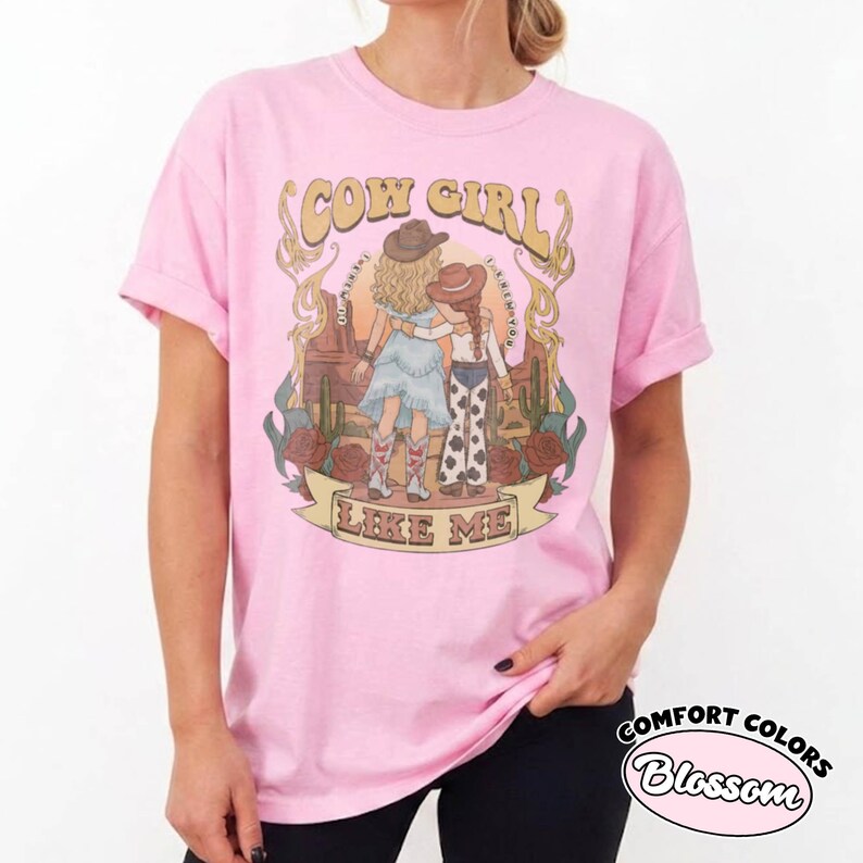 May include: A light pink t-shirt featuring a graphic print of two figures in cowboy hats and boots standing in a desert landscape with cacti and roses. The text COW GIRL and LIKE ME is printed in a decorative font above and below the illustration.