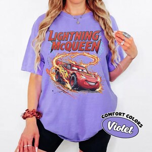 Puede incluir: Una camiseta de color violeta con un dise&ntilde;o de Lightning McQueen de la pel&iacute;cula Cars. El dise&ntilde;o incluye el texto "LIGHTNING McQUEEN" en rojo y amarillo, con el coche rodeado de llamas. La camiseta tambi&eacute;n tiene el texto "COMFORT COLORS" y "Violet".