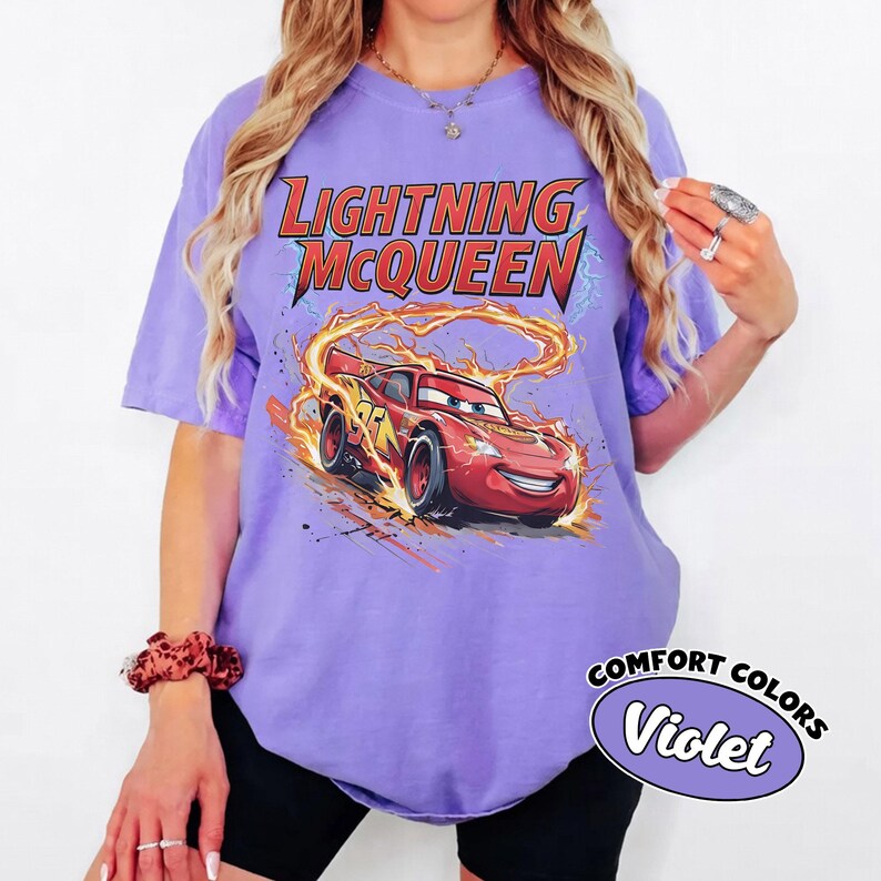 Puede incluir: Una camiseta de color violeta con un dise&ntilde;o de Lightning McQueen de la pel&iacute;cula Cars. El dise&ntilde;o incluye el texto "LIGHTNING McQUEEN" en rojo y amarillo, con el coche rodeado de llamas. La camiseta tambi&eacute;n tiene el texto "COMFORT COLORS" y "Violet".