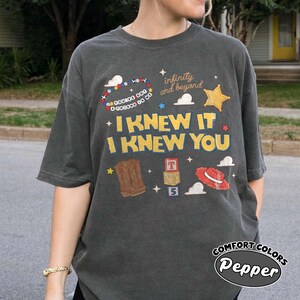 May include: A charcoal grey short-sleeve t-shirt featuring a graphic print with the text "I KNEW IT I KNEW YOU", "infinity and beyond", and "YOU'VE GOT A FRIEND IN ME". The design includes illustrations of a cowboy boot, a red hat, and blocks.