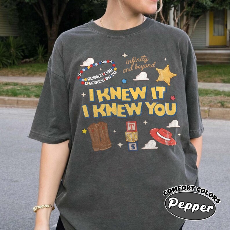 May include: A charcoal grey short-sleeve t-shirt featuring a graphic print with the text "I KNEW IT I KNEW YOU", "infinity and beyond", and "YOU'VE GOT A FRIEND IN ME". The design includes illustrations of a cowboy boot, a red hat, and blocks.