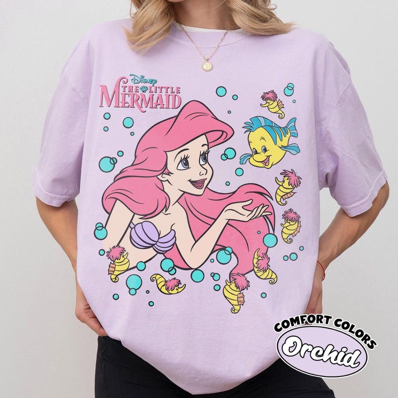 Puede incluir: Una camiseta de manga corta de color morado claro con un estampado gr&aacute;fico de Ariel de La Sirenita con el pelo largo y rosa, un top de conchas morado, el pez Flounder y varios caballitos de mar rodeados de burbujas azules y el t&iacute;tulo.