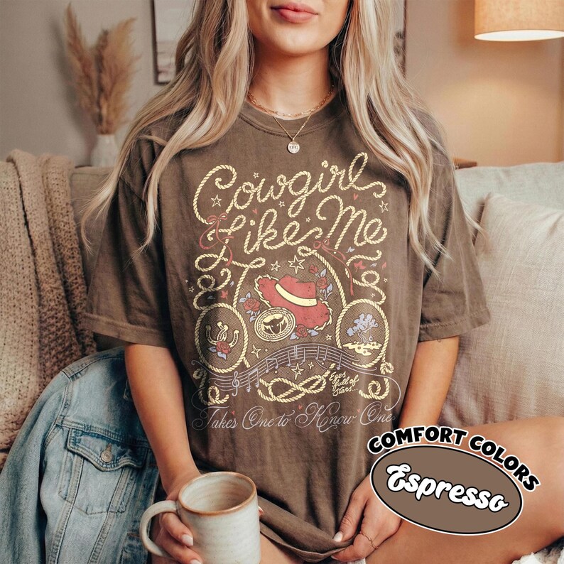 以下が含まれることがあります： Cowgirl Like Meと書かれたロープ風の文字が特徴のダークブラウンの半袖Tシャツ。プリントには赤いカウボーイハット、蹄鉄、音符が含まれています。下部にはTakes One to Know Oneというテキストがデザインされています。