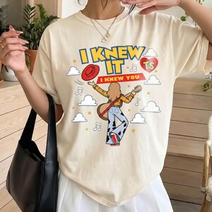 May include: A cream-coloured short-sleeve t-shirt featuring a graphic print with the text I KNEW IT and I KNEW YOU. The design includes a person playing a guitar, a red hat, a red heart with TS, musical notes, clouds, and yellow stars.