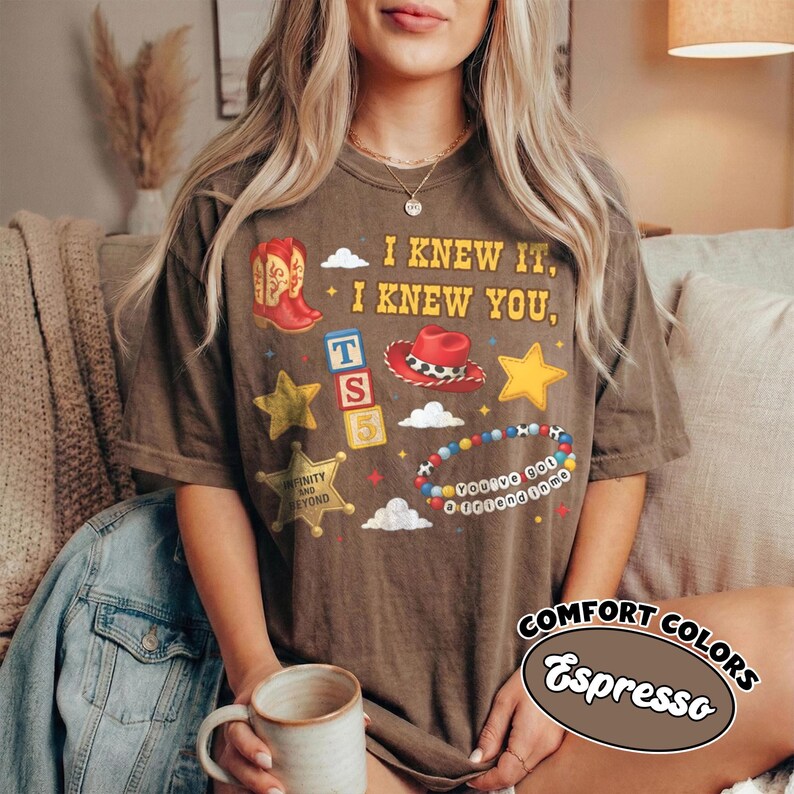 Peut inclure: Un t-shirt &agrave; manches courtes marron fonc&eacute; avec une impression graphique comprenant le texte I KNEW IT, I KNEW YOU, une botte de cowboy rouge, un chapeau de cowboy rouge, des &eacute;toiles jaunes, une &eacute;toile de sh&eacute;rif, des blocs color&eacute;s.