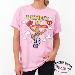 May include: A light pink t-shirt featuring a graphic of a person with long hair playing an acoustic guitar, wearing a yellow plaid shirt and cow-print bell-bottom trousers. The text reads I KNEW IT I KNEW YOU with a red hat, heart, and clouds.