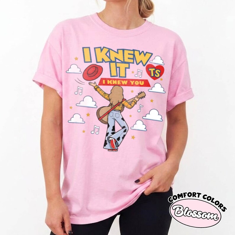 May include: A light pink t-shirt featuring a graphic of a person with long hair playing an acoustic guitar, wearing a yellow plaid shirt and cow-print bell-bottom trousers. The text reads I KNEW IT I KNEW YOU with a red hat, heart, and clouds.