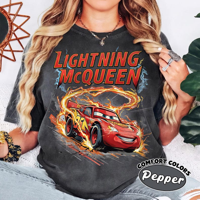 Puede incluir: Camiseta gris oscuro con Lightning McQueen de la pel&iacute;cula Cars. El gr&aacute;fico muestra el coche de carreras rojo con llamas y el texto "Lightning McQueen". La camiseta est&aacute; etiquetada como "Comfort Colors Pepper".