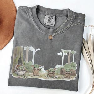 Puede incluir: Camiseta gris Comfort Colors con una escena de dibujos animados con varios personajes de Grogu en un entorno forestal. La camiseta tiene cuello redondo y mangas cortas. Un collar plateado con un colgante est&aacute; sobre la camiseta.