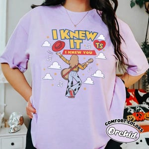 May include: A light purple t-shirt featuring a graphic print of a person with long hair playing a guitar, wearing a yellow plaid shirt and cow-print trousers. The text reads I KNEW IT and I KNEW YOU, accompanied by a red hat, a red heart, and clouds.