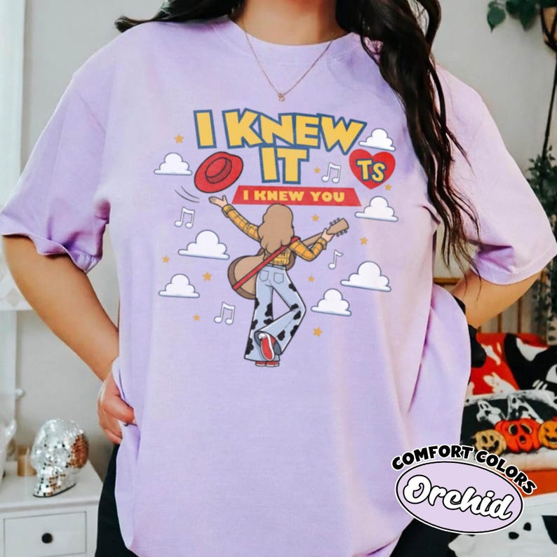 May include: A light purple t-shirt featuring a graphic print of a person with long hair playing a guitar, wearing a yellow plaid shirt and cow-print trousers. The text reads I KNEW IT and I KNEW YOU, accompanied by a red hat, a red heart, and clouds.