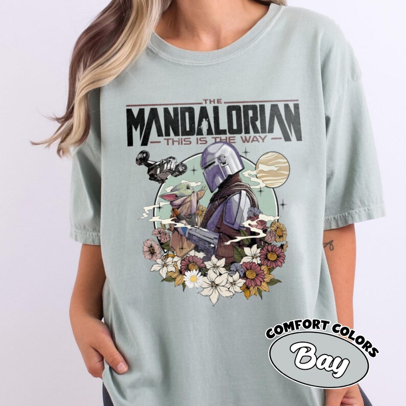 Op de afbeelding: Een licht saliegroen t-shirt met een grafische print van The Mandalorian en Grogu. Het ontwerp bevat de tekst THE MANDALORIAN THIS IS THE WAY, een ruimteschip, een planeet en een groep kleurrijke bloemen rondom de centrale personages.