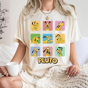 May include: A cream-coloured t-shirt displaying nine squares, each with a different Pluto cartoon character illustration. The word "PLUTO" is printed in yellow below. The t-shirt is being worn by a person.