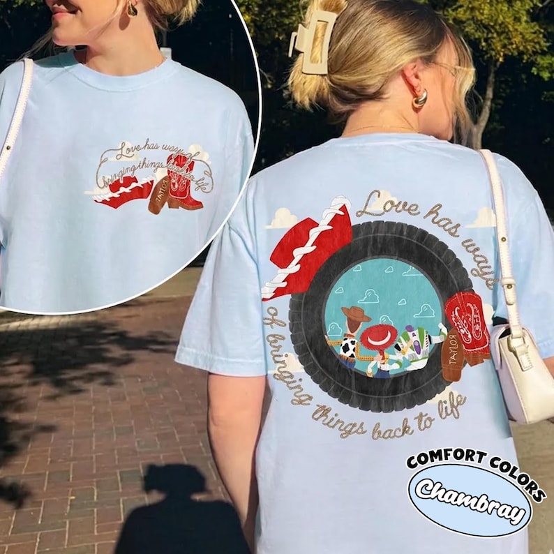 May include: A light blue t-shirt featuring a graphic on the back and a smaller version on the front. The design includes a black tire framing a scene with cartoon characters, a red cowboy hat, and red boots with the name Taylor, plus the text Love has ways of bringing things back to life.