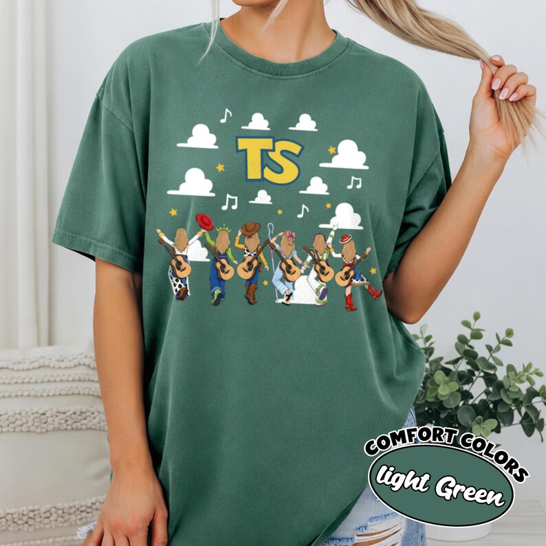Może przedstawiać: Leśno-zielony t-shirt z kr&oacute;tkim rękawem z grafiką przedstawiającą sześć postaci z gitarami, białymi chmurami, nutami i ż&oacute;łtymi gwiazdkami. Litery TS są nadrukowane na ż&oacute;łto z niebieskim obrysem nad postaciami. Koszulka ma okrągły dekolt.