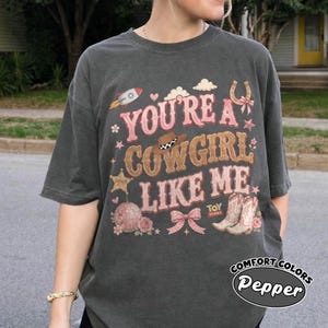 May include: A charcoal grey short-sleeve t-shirt featuring a graphic print with the text "YOU'RE A COWGIRL LIKE ME" in pink and tan. The design includes a rocket, a cowboy hat, a sheriff star, a disco ball, a bow, and a pair of cowboy boots.