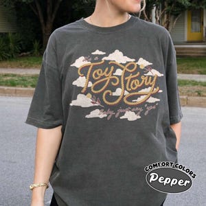 May include: A charcoal gray short-sleeve t-shirt featuring a graphic print of white clouds and the text "it's a Toy Story" written in a yellow rope-style font, with the additional text "baby, just say 'yes'" printed below in a cursive script.