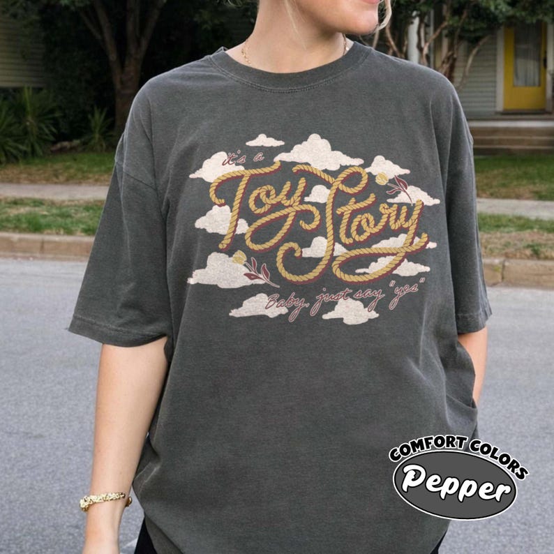 May include: A charcoal gray short-sleeve t-shirt featuring a graphic print of white clouds and the text "it's a Toy Story" written in a yellow rope-style font, with the additional text "baby, just say 'yes'" printed below in a cursive script.