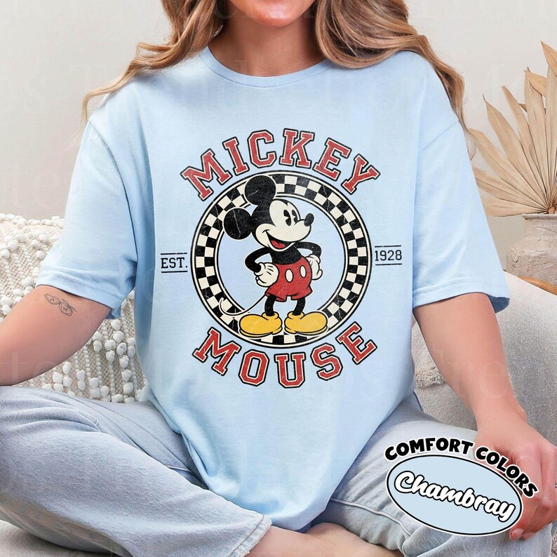 May include: Light blue t-shirt featuring a distressed Mickey Mouse graphic. The design includes the words "Mickey Mouse" in a vintage font, with "EST. 1928" and a checkered border. The shirt also has the text "COMFORT COLORS Chambray".