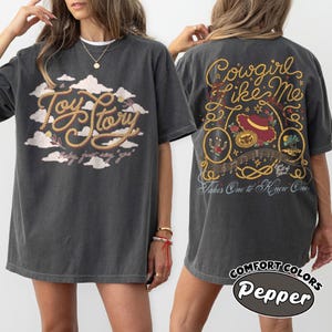 May include: A charcoal gray t-shirt featuring a front graphic of clouds and the text Toy Story with the phrase baby, just say yes. The back graphic displays a rope design, a hat, and the text Cowgirl Like Me, Takes One to Know One.