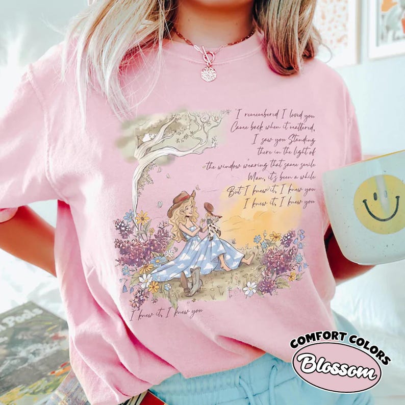May include: A light pink t-shirt featuring a graphic of a person in a blue dress and cowboy hat sitting in a field of flowers under a tree. The text reads: I remembered I loved you, Came back when it mattered, I saw you standing there in the light of the window wearing that same smile, Man, it's been a while, But I knew it, I knew it, I knew it, I knew you.