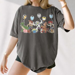May include: Dark grey t-shirt with a colourful graphic of Toy Story characters holding balloons. The design includes Woody, Buzz Lightyear, Jessie, and other characters from the animated film. The shirt has a relaxed fit.