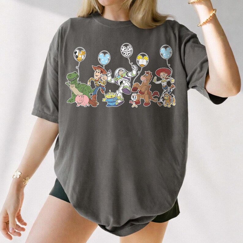 May include: Dark grey t-shirt with a colourful graphic of Toy Story characters holding balloons. The design includes Woody, Buzz Lightyear, Jessie, and other characters from the animated film. The shirt has a relaxed fit.