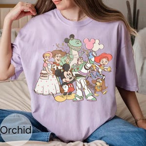 May include: A lilac t-shirt with a graphic of cartoon characters such as Mickey Mouse, Woody, and Buzz Lightyear. The shirt has a crew neck and short sleeves. The word "Orchid" is printed at the bottom of the shirt.