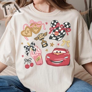 May include: Cream-colored t-shirt featuring a colorful graphic design. The design includes a red race car, a checkered flag, a milkshake, and heart-shaped cookies with text. Additional elements include stars and bows.