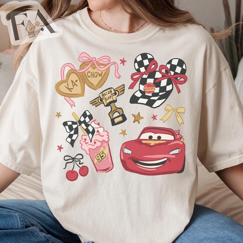 May include: Cream-colored t-shirt featuring a colorful graphic design. The design includes a red race car, a checkered flag, a milkshake, and heart-shaped cookies with text. Additional elements include stars and bows.