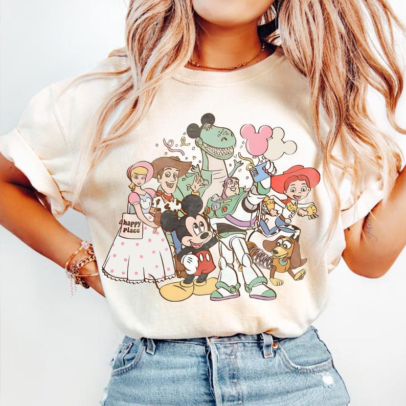 May include: Cream-coloured t-shirt with a colourful graphic of Disney characters, including Mickey Mouse, Woody, Buzz Lightyear, and others. The phrase "happy place" is printed on the shirt. The shirt is paired with blue denim jeans.
