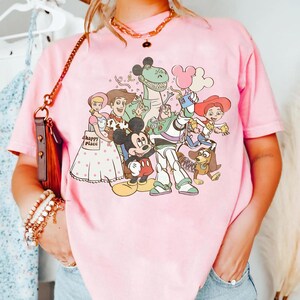 May include: Pale pink t-shirt with a cartoon graphic of various characters, including Mickey Mouse, Woody, Buzz Lightyear, and others. The shirt has a crew neck and short sleeves. The text "happy place" is visible on the shirt.
