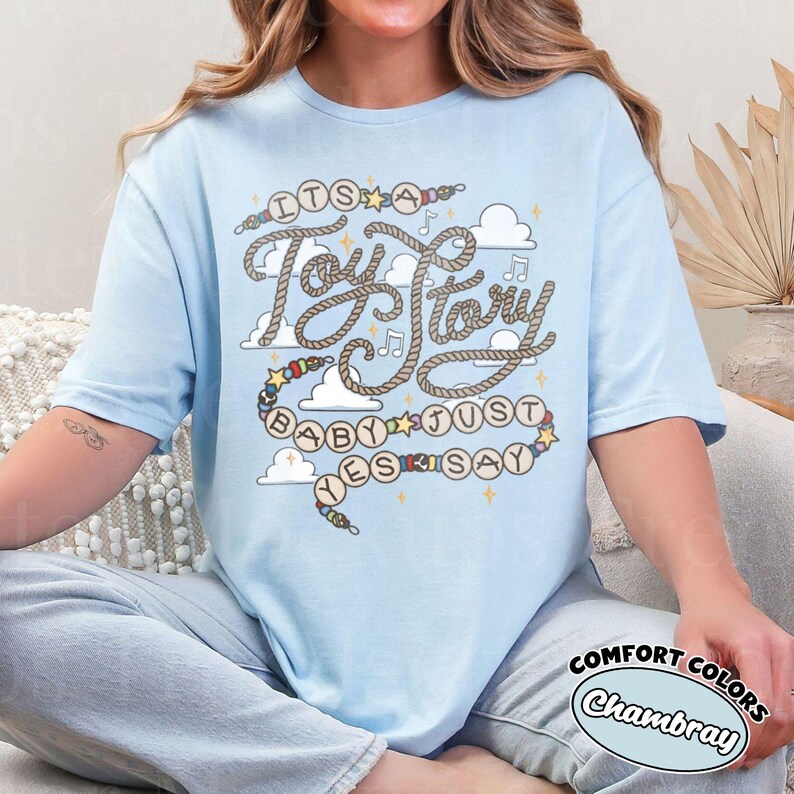 May include: A light blue t-shirt featuring a graphic design with the text "IT'S A Toy Story BABY JUST YES SAY". The design includes rope-style lettering, colorful beads, musical notes, and white clouds against a light blue fabric background.