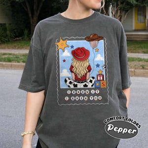 May include: A charcoal gray t-shirt featuring a graphic of a blonde person wearing a red cowboy hat, sitting with their back to the viewer. The design includes a brown cowboy hat balloon, a yellow star, alphabet blocks, and the text I KNEW IT I KNEW YOU.