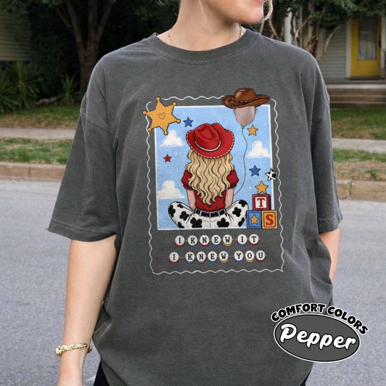 May include: A charcoal gray t-shirt featuring a graphic of a blonde person wearing a red cowboy hat, sitting with their back to the viewer. The design includes a brown cowboy hat balloon, a yellow star, alphabet blocks, and the text I KNEW IT I KNEW YOU.