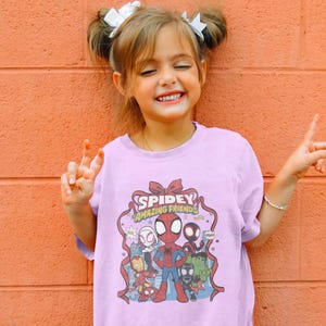May include: A light purple t-shirt featuring the text "Spidey Amazing Friends" with cartoon characters. The shirt is worn by a young person with white bows in their hair, smiling and making a peace sign.