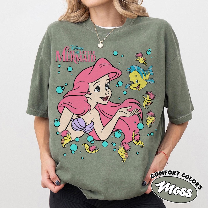 Puede incluir: Una camiseta verde oliva con un gr&aacute;fico de Ariel de La Sirenita con cabello rosa, un top de conchas morado y el pez Flounder, rodeados de peque&ntilde;os caballitos de mar amarillos y burbujas azul cerceta, con el logo de Disney La Sirenita.
