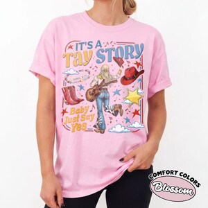 May include: A light pink t-shirt featuring a graphic print with the text IT'S A TAY STORY and Baby Just Say Yes. The design includes a person playing an acoustic guitar, red cowboy boots, a red cowboy hat, a yellow star, clouds, and musical notes.