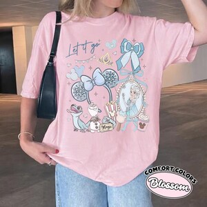 May include: A light pink t-shirt featuring a graphic print with the text "Let it go" and "Magic". The design includes a character in a mirror, a blue bow, mouse ears, a lizard, a snowman, popcorn, and a drink cup with a mouse-shaped lid.