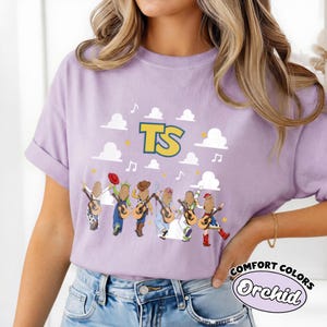 Może przedstawiać: Jasnofioletowy t-shirt z kr&oacute;tkim rękawem z nadrukiem graficznym przedstawiającym sześć postaci z kresk&oacute;wek widzianych od tyłu, z kt&oacute;rych każda trzyma gitarę. Powyżej znajdują się białe chmury, ż&oacute;łte gwiazdy, nuty oraz duże ż&oacute;łte litery TS.
