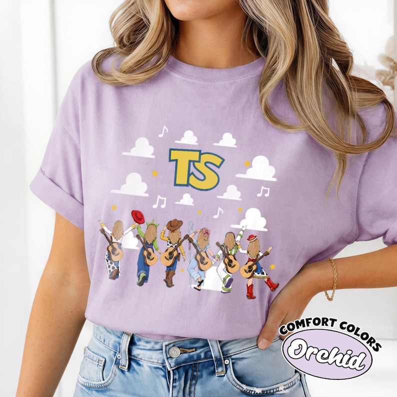 Może przedstawiać: Jasnofioletowy t-shirt z kr&oacute;tkim rękawem z nadrukiem graficznym przedstawiającym sześć postaci z kresk&oacute;wek widzianych od tyłu, z kt&oacute;rych każda trzyma gitarę. Powyżej znajdują się białe chmury, ż&oacute;łte gwiazdy, nuty oraz duże ż&oacute;łte litery TS.
